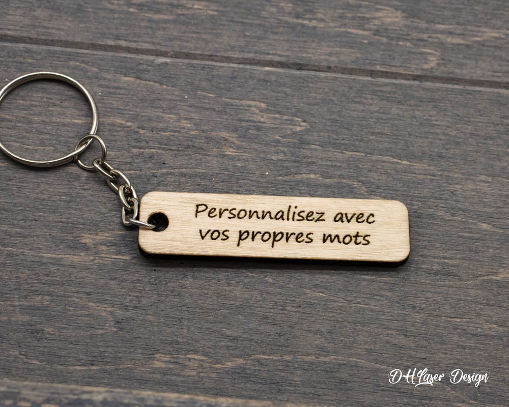 Porte clé personnalisable en bois clair DHLaser Design avec texte gravé au laser posé sur planche de bois brut foncé et anneau métallique argenté